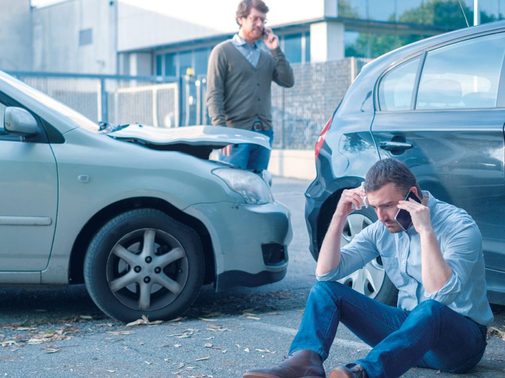 motorista sentado no chão falando ao telefone, e o seu carro acidentado no fundo 